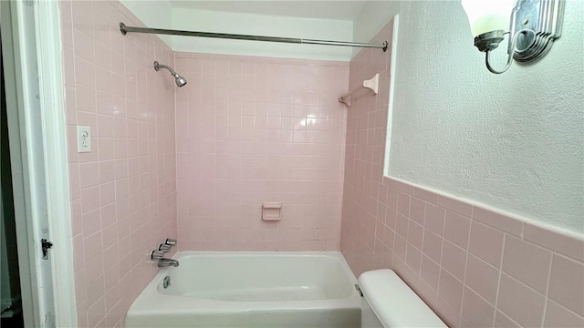 bathroom with a textured wall, toilet, wainscoting, washtub / shower combination, and tile walls