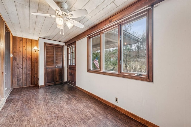 empty room featuring a ceiling fan, wooden walls, baseboards, and dark wood-type flooring