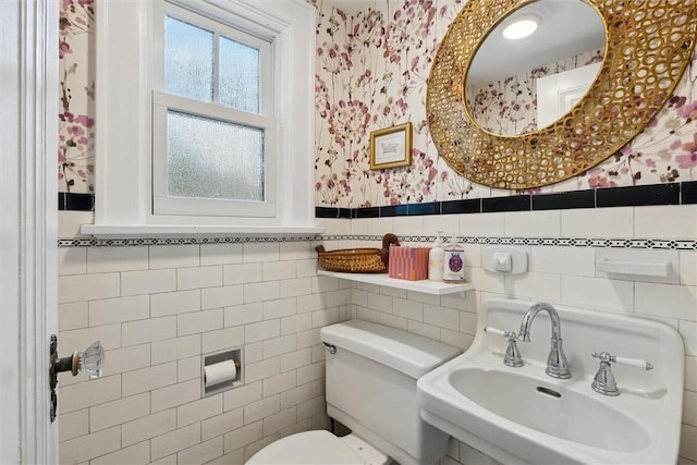 bathroom featuring wallpapered walls, a wainscoted wall, toilet, tile walls, and a sink