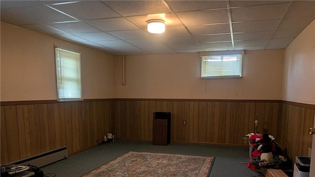 basement with wooden walls, carpet floors, wainscoting, baseboard heating, and a paneled ceiling