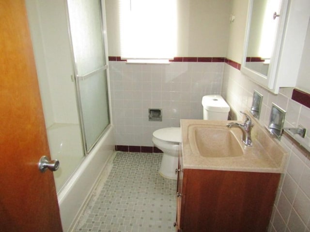 bathroom featuring toilet, a wainscoted wall, vanity, tile walls, and combined bath / shower with glass door