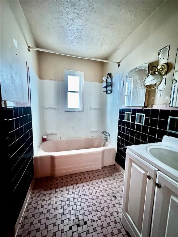 full bath with wainscoting,  shower combination, a textured ceiling, vanity, and tile walls