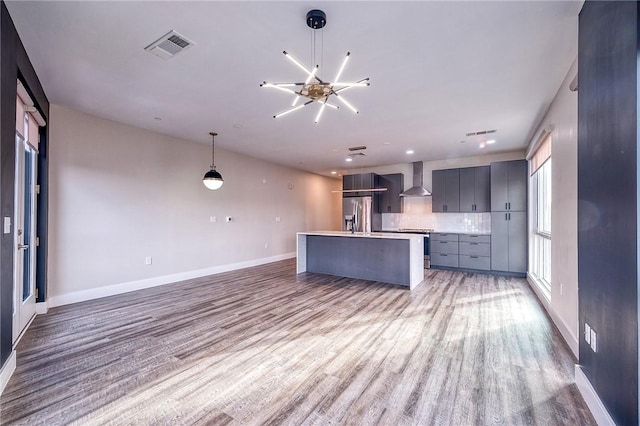 kitchen with light wood-type flooring, wall chimney range hood, a center island with sink, and appliances with stainless steel finishes