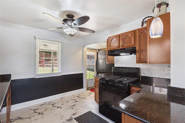 kitchen with brown cabinets, dark stone countertops, marble finish floor, black gas stove, and under cabinet range hood