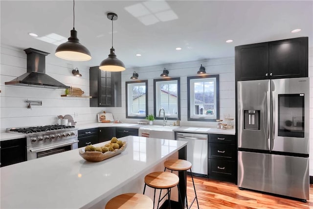 kitchen with stainless steel appliances, light countertops, a sink, wall chimney range hood, and dark cabinets