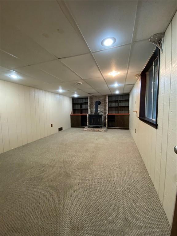 finished basement featuring a wood stove, carpet, wooden walls, and a drop ceiling