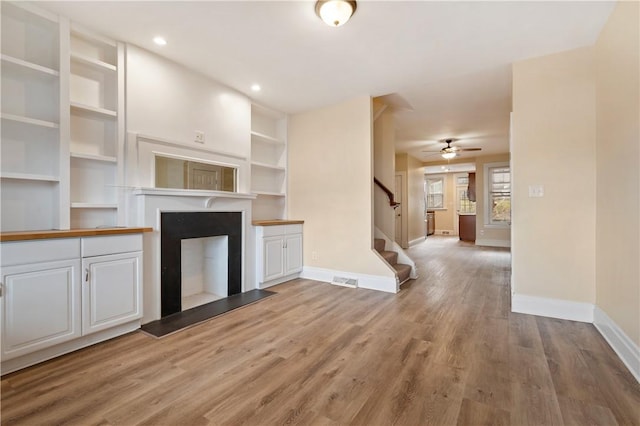 unfurnished living room with a fireplace with raised hearth, light wood-style flooring, baseboards, and stairs