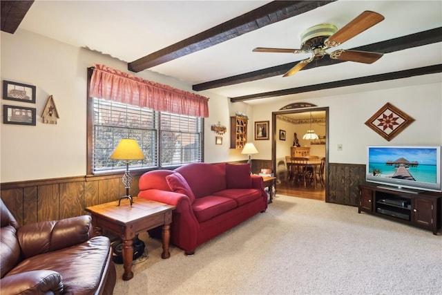 carpeted living room with wood walls, a ceiling fan, beam ceiling, and wainscoting