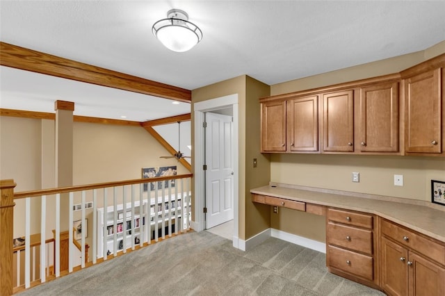 unfurnished office featuring beam ceiling, built in desk, light colored carpet, a ceiling fan, and baseboards