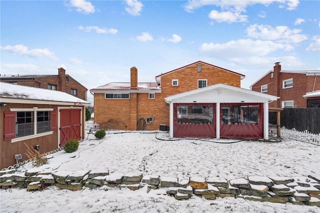 snow covered back of property featuring an outbuilding, brick siding, a chimney, a sunroom, and fence