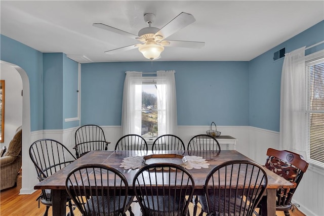 dining room with visible vents, arched walkways, a ceiling fan, a wainscoted wall, and light wood-type flooring