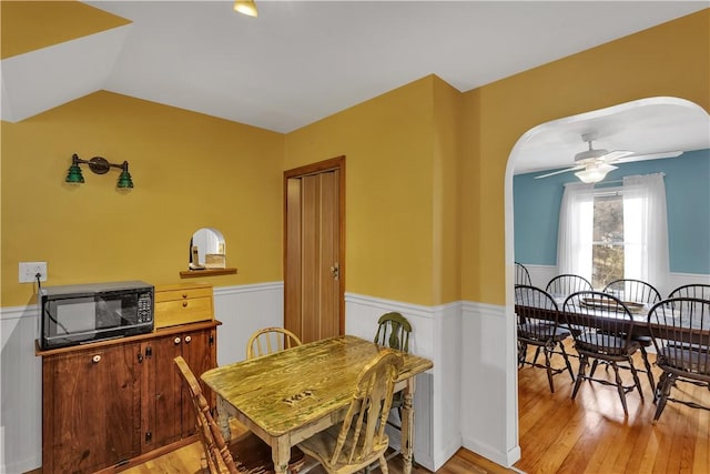 dining area with arched walkways, light wood-style flooring, a wainscoted wall, and a ceiling fan