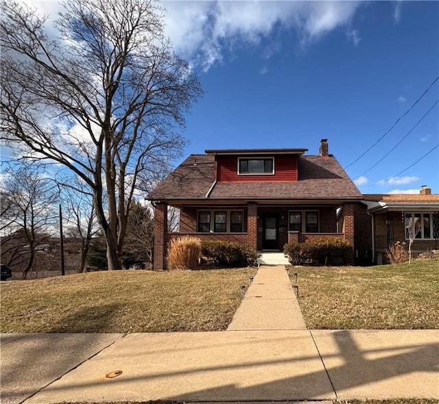 view of front of property with brick siding, a shingled roof, a front lawn, a porch, and a chimney