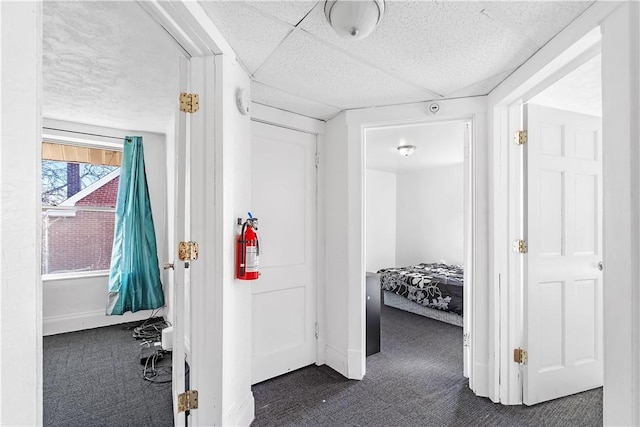 hall with baseboards, dark colored carpet, and a drop ceiling