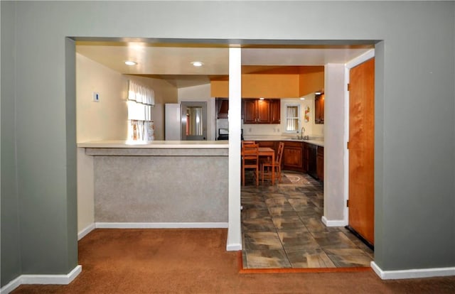 kitchen with a sink, baseboards, light countertops, brown cabinetry, and dark carpet