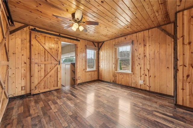 empty room featuring a barn door, dark wood-type flooring, wood ceiling, ceiling fan, and wooden walls