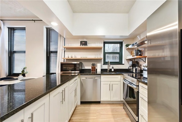 kitchen with a sink, appliances with stainless steel finishes, light wood-type flooring, open shelves, and dark stone countertops