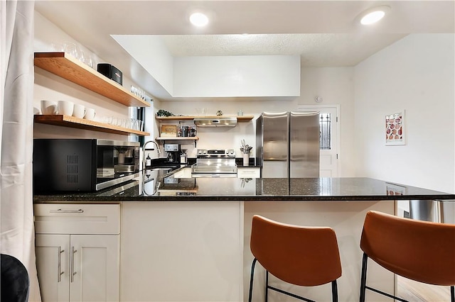 kitchen with under cabinet range hood, stainless steel appliances, a breakfast bar, a sink, and open shelves