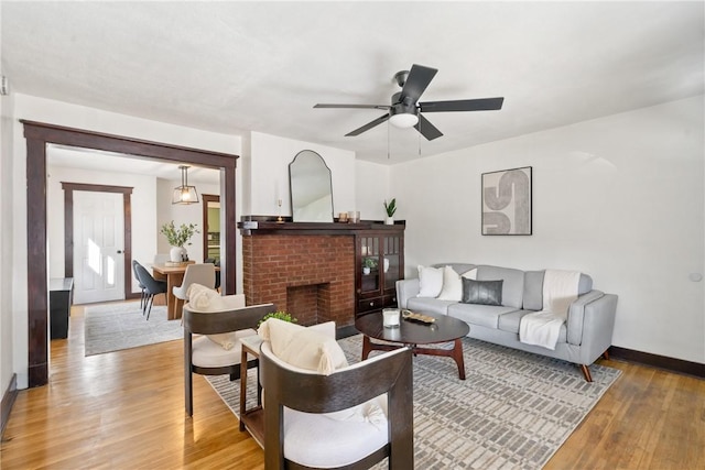 living area featuring baseboards, light wood-type flooring, a ceiling fan, and a brick fireplace