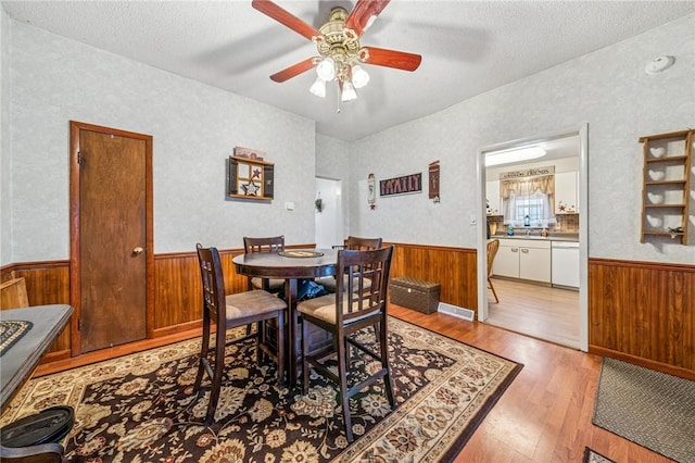 dining space with ceiling fan, a wainscoted wall, wood walls, light wood-style flooring, and a textured ceiling