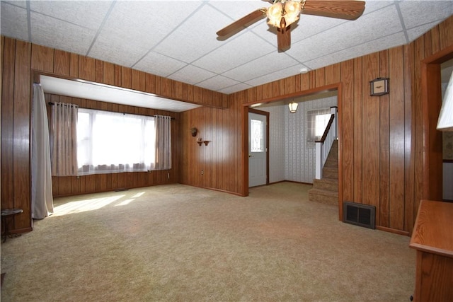 unfurnished living room featuring wooden walls, carpet, visible vents, ceiling fan, and stairs