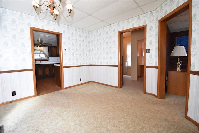 carpeted empty room featuring a drop ceiling, a wainscoted wall, an inviting chandelier, and wallpapered walls