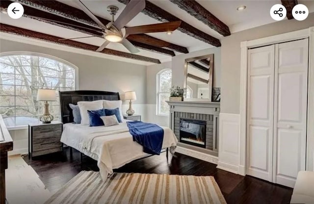 bedroom featuring dark wood finished floors, a fireplace, ceiling fan, wainscoting, and beamed ceiling