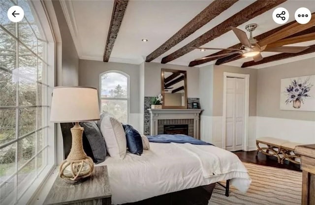 bedroom with beam ceiling, wainscoting, a fireplace, wood finished floors, and a ceiling fan