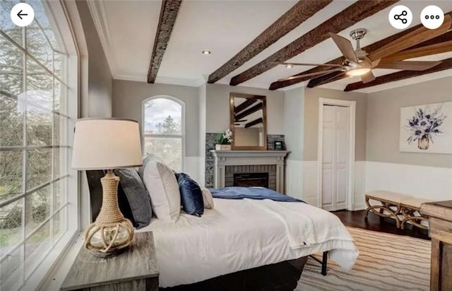 bedroom featuring a ceiling fan, wood finished floors, beam ceiling, wainscoting, and a brick fireplace