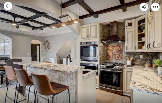 kitchen with cream cabinetry, coffered ceiling, stainless steel appliances, wall chimney range hood, and decorative backsplash