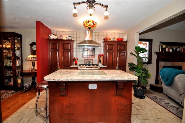 kitchen featuring wall chimney exhaust hood, a center island, dark brown cabinets, light stone counters, and a breakfast bar area