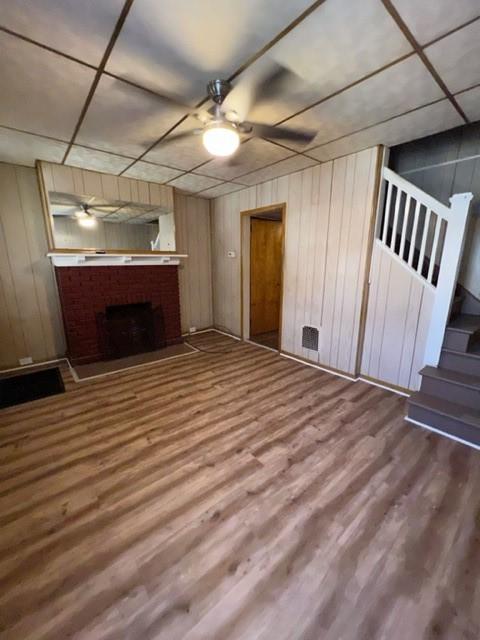 unfurnished living room featuring a ceiling fan, wood finished floors, wood walls, a fireplace, and stairs