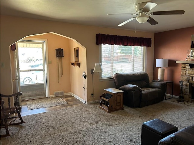 carpeted living room with visible vents, a ceiling fan, arched walkways, a stone fireplace, and baseboards