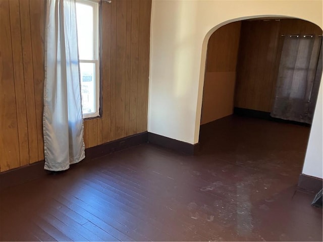 unfurnished room featuring baseboards, arched walkways, wooden walls, and dark wood-style flooring