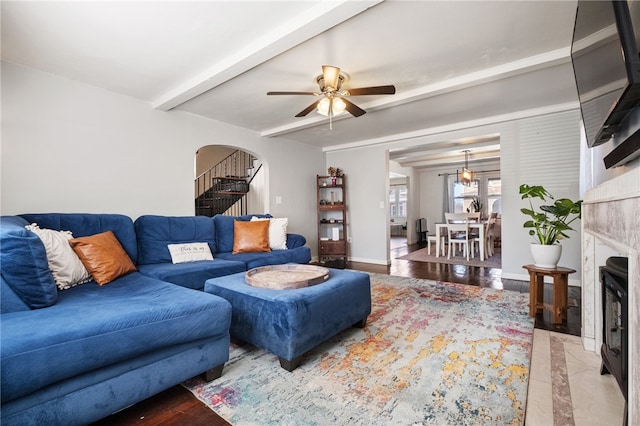 living area with beamed ceiling, a fireplace with flush hearth, stairs, arched walkways, and a ceiling fan