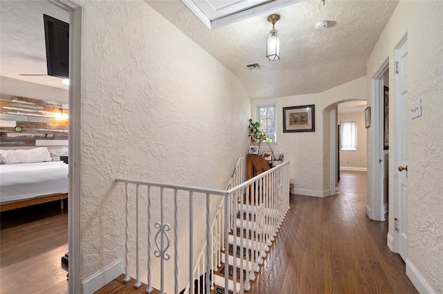 hall with visible vents, a textured wall, an upstairs landing, wood finished floors, and arched walkways