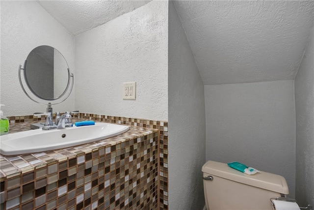 half bath with lofted ceiling, a sink, a textured ceiling, toilet, and a textured wall