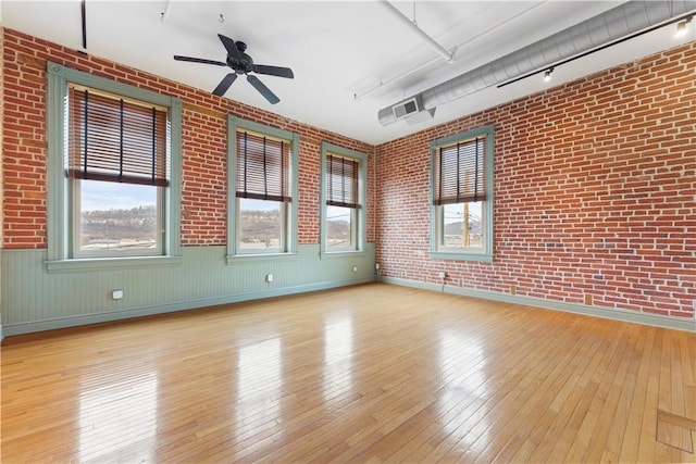 empty room featuring brick wall, wainscoting, rail lighting, a ceiling fan, and wood-type flooring