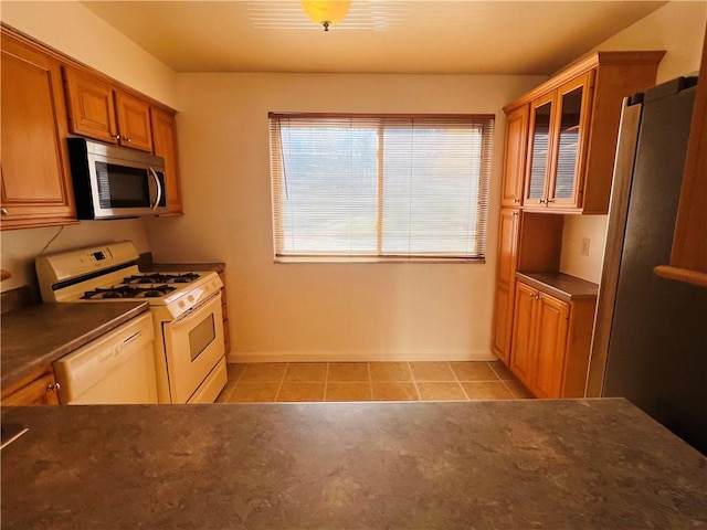 kitchen featuring dark countertops, white appliances, light tile patterned flooring, brown cabinetry, and glass insert cabinets