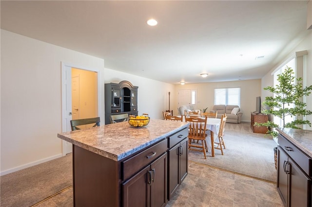 kitchen featuring dark brown cabinetry, light colored carpet, a kitchen island, and baseboards