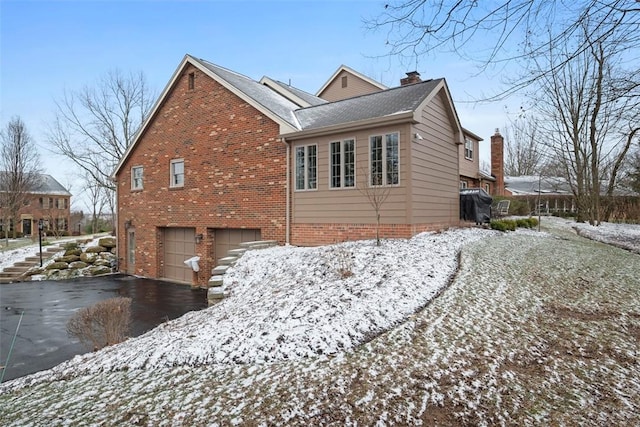 view of snow covered exterior with aphalt driveway, a garage, a chimney, and brick siding