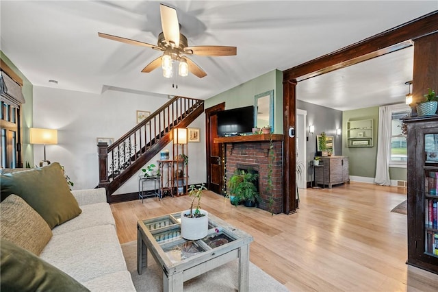 living room featuring a ceiling fan, wood finished floors, baseboards, a brick fireplace, and stairs