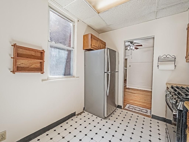 kitchen featuring black gas stove, a drop ceiling, light floors, freestanding refrigerator, and brown cabinetry