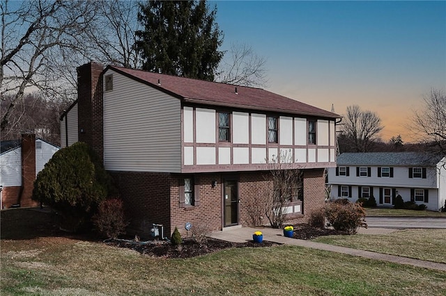 back of house at dusk with brick siding, a chimney, and a yard