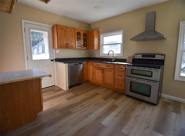 kitchen with brown cabinetry, a sink, appliances with stainless steel finishes, wall chimney exhaust hood, and light wood-type flooring