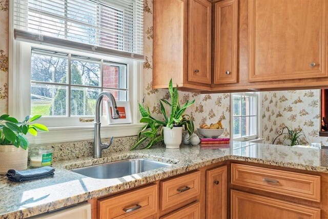 kitchen featuring wallpapered walls, brown cabinetry, light stone countertops, and a sink