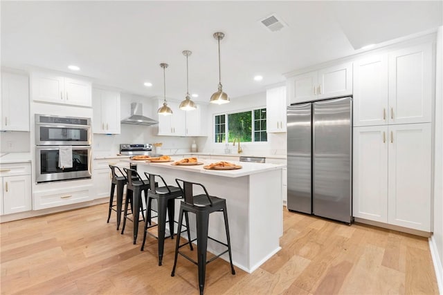 kitchen with a breakfast bar, stainless steel appliances, light wood-style floors, wall chimney exhaust hood, and light countertops