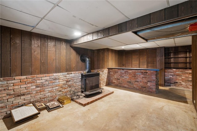 finished basement with a paneled ceiling, brick wall, wood walls, and a wood stove