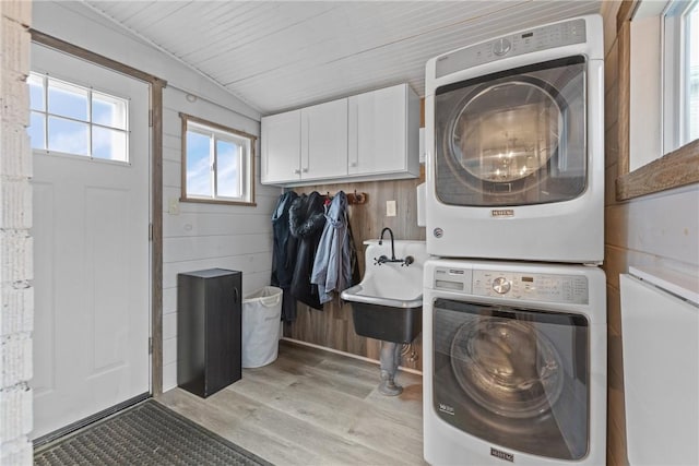 laundry area featuring a sink, cabinet space, wood walls, light wood finished floors, and stacked washer / dryer