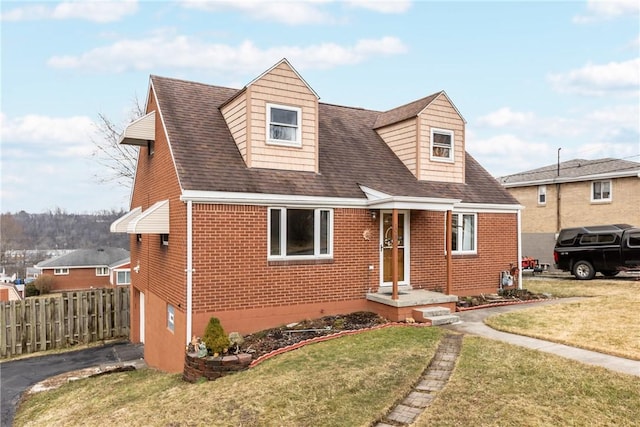 new england style home with brick siding, roof with shingles, a front yard, and fence